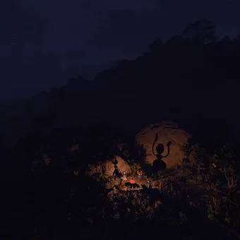 Big Walk at night: a dark bushland ridge under a starry sky, with a small firelit clearing and figures gathered around it in the distance (May 2025 build)