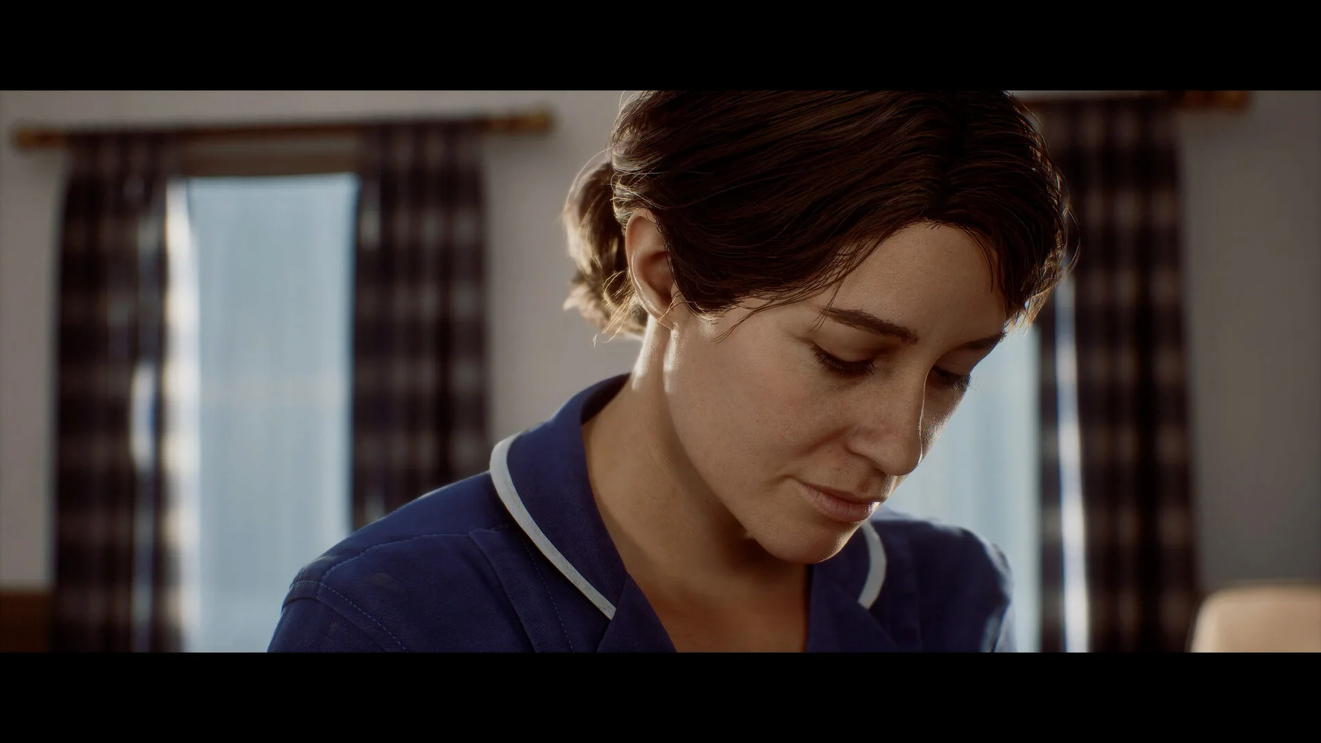 A woman in a blue clinic uniform looking down, window light behind her.